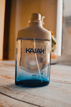 A glass bottle with a beige cap and a transparent blue lower section, featuring the brand name 'KAIAK' printed in blue. The bottle is positioned on a light wooden surface with blurred background elements visible through a window.
