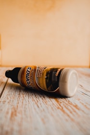 A plastic bottle with a black cap lying on its side on a wooden surface. The label reads 'Aroma's Mantenimiento' and features an image of an interior space. The background appears to be a light-colored wall.