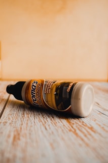 A plastic bottle with a black cap lying on its side on a wooden surface. The label reads 'Aroma's Mantenimiento' and features an image of an interior space. The background appears to be a light-colored wall.