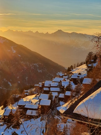 A picturesque alpine village with snow-covered rooftops set on a mountainside during sunset. The sky is clear with a golden hue, and the distant mountain range is visible under the warm, glowing light of the sun.