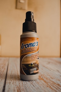 A bottle of air freshener labeled 'Aroma's Antitabaco' placed on a wooden surface with a light beige background. The label also mentions eliminating strong odors for a longer time.
