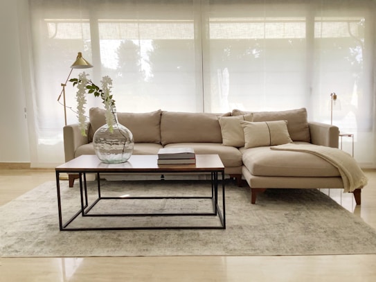 A modern living room setup features a beige sectional sofa adorned with matching cushions and a light blanket draped on one side. In front of the sofa is a sleek wooden coffee table with a metal frame, supporting a large glass vase with decorative branches and a couple of stacked books. The room is well-lit with natural light filtering through large sheer curtains, creating an airy and tranquil atmosphere. A minimalist floor lamp with a gold finish stands nearby, complementing the sophisticated and elegant setting.