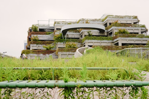 A multi-level structure covered in green plants and grass, with a modern and urban design. The foreground has lush vegetation, and there are pathways leading up to the building, creating a harmonious blend with nature.