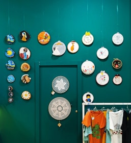Several colorful embroidery hoops and decorative items are displayed on a teal wall. The arrangement includes various designs, such as abstract patterns, floral motifs, and figures. Below, a rack holds several brightly colored t-shirts hanging side by side.