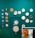 Several colorful embroidery hoops and decorative items are displayed on a teal wall. The arrangement includes various designs, such as abstract patterns, floral motifs, and figures. Below, a rack holds several brightly colored t-shirts hanging side by side.