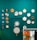 Several colorful embroidery hoops and decorative items are displayed on a teal wall. The arrangement includes various designs, such as abstract patterns, floral motifs, and figures. Below, a rack holds several brightly colored t-shirts hanging side by side.