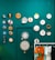 Several colorful embroidery hoops and decorative items are displayed on a teal wall. The arrangement includes various designs, such as abstract patterns, floral motifs, and figures. Below, a rack holds several brightly colored t-shirts hanging side by side.