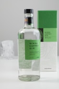 A bottle of Nikka Coffey Gin with a green label and a black cap is seen in the foreground. In the background, there is the box packaging that also has a green and white design matching the bottle's label. The setting appears to be minimalistic with a clean, light-colored background.