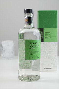 A bottle of Nikka Coffey Gin with a green label and a black cap is seen in the foreground. In the background, there is the box packaging that also has a green and white design matching the bottle's label. The setting appears to be minimalistic with a clean, light-colored background.