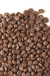 A large pile of roasted coffee beans spread across a flat surface. The beans are uniformly dark brown with a glossy appearance, showcasing the distinct lines and curves typical of coffee beans.