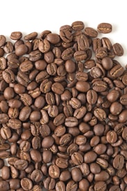 A large pile of roasted coffee beans spread across a flat surface. The beans are uniformly dark brown with a glossy appearance, showcasing the distinct lines and curves typical of coffee beans.
