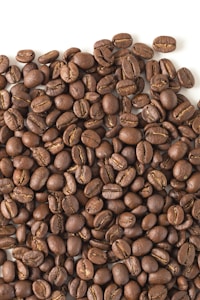 A large pile of roasted coffee beans spread across a flat surface. The beans are uniformly dark brown with a glossy appearance, showcasing the distinct lines and curves typical of coffee beans.