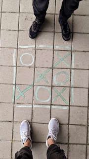 A game of tic-tac-toe is drawn on a tiled sidewalk with chalk, showing alternating X and O symbols. Two pairs of feet are visible, one in black shoes and the other in white sneakers, positioned at opposite ends of the grid.