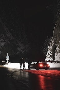 A snowy mountain road at night is dimly lit, revealing a stopped car with its tail lights casting a red glow on the road. Three people are standing near the car, silhouettes against the backdrop of snow-covered rock walls and forest.