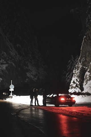 A snowy mountain road at night is dimly lit, revealing a stopped car with its tail lights casting a red glow on the road. Three people are standing near the car, silhouettes against the backdrop of snow-covered rock walls and forest.