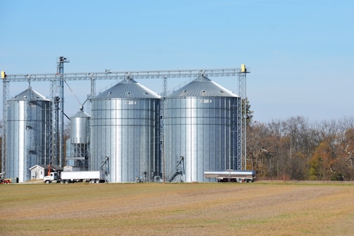 Several large metal grain silos stand on a farm field with a clear blue sky above. A semi-truck and another vehicle are parked nearby, suggesting agricultural activity. Trees with autumn foliage are visible in the background.