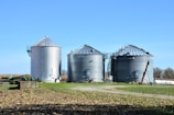Three large metallic silos stand prominently in a field with clear blue skies overhead. The silos appear to be used for grain storage and are surrounded by a combination of grass and fallen leaves. To the left, a farming implement is partially visible. In the background, some trees and vegetation can be seen.