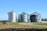 Three large metallic silos stand prominently in a field with clear blue skies overhead. The silos appear to be used for grain storage and are surrounded by a combination of grass and fallen leaves. To the left, a farming implement is partially visible. In the background, some trees and vegetation can be seen.