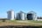 Three large metallic silos stand prominently in a field with clear blue skies overhead. The silos appear to be used for grain storage and are surrounded by a combination of grass and fallen leaves. To the left, a farming implement is partially visible. In the background, some trees and vegetation can be seen.