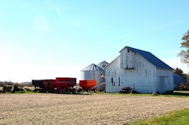 A rural farm scene with a weathered white barn and agricultural equipment, including two large red wagons and a silo, situated on a field under a clear blue sky.