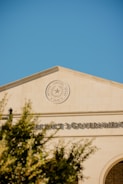 A government building with a prominent seal that reads 'Harris County, Texas' and 'Since 1846' is featured. The architecture is classic with a clean, tan facade and large text displaying 'PRECINCT 3 GOVERNMENT'. The day is clear with a bright blue sky and a few blurred tree branches in the foreground.