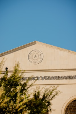 A government building with a prominent seal that reads 'Harris County, Texas' and 'Since 1846' is featured. The architecture is classic with a clean, tan facade and large text displaying 'PRECINCT 3 GOVERNMENT'. The day is clear with a bright blue sky and a few blurred tree branches in the foreground.