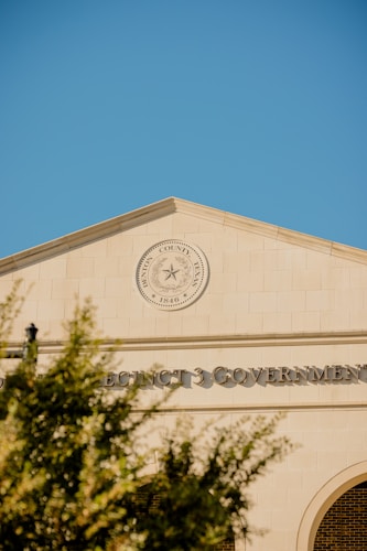 A government building with a prominent seal that reads 'Harris County, Texas' and 'Since 1846' is featured. The architecture is classic with a clean, tan facade and large text displaying 'PRECINCT 3 GOVERNMENT'. The day is clear with a bright blue sky and a few blurred tree branches in the foreground.