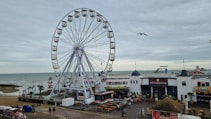 A large Ferris wheel stands prominently on a pier by the sea, with cloudy skies overhead. Below the wheel, there are various buildings and attractions, including eateries and shops. The sandy beach and a lighthouse are visible in the background, along with some people walking around. Seagulls are flying above the area, adding to the seaside atmosphere.