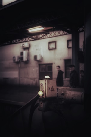 An industrial setting features a dimly lit area with a control box or panel in the foreground, illuminated by a small light. In the background, a blurred view of people passing by a building wall with mounted equipment and small windows can be seen.