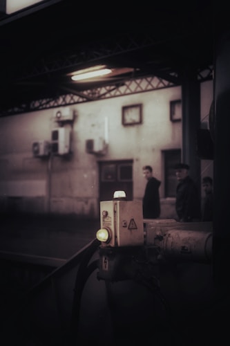 An industrial setting features a dimly lit area with a control box or panel in the foreground, illuminated by a small light. In the background, a blurred view of people passing by a building wall with mounted equipment and small windows can be seen.