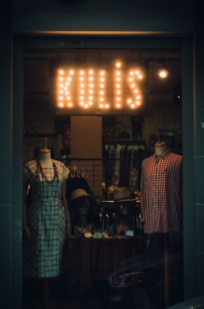 A dimly lit shop window display showcases two mannequins dressed in vintage-style clothing. The left mannequin wears a checked dress, while the right one sports a red and white plaid shirt. Accessories like hats and jewelry are arranged on a table in front of the mannequins. Above them, a sign with the word 'KULIŞ' is illuminated with retro-style light bulbs, creating a warm glow.