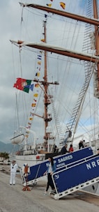 A large sailing ship with tall masts is docked at a port. The masts are adorned with colorful nautical flags, and a Portuguese flag is prominently displayed. People are descending a gangway labeled 'NRP SAGRES PORTUGAL'. A person dressed in a sailor's uniform stands nearby on the dock.