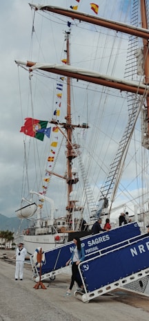 A large sailing ship with tall masts is docked at a port. The masts are adorned with colorful nautical flags, and a Portuguese flag is prominently displayed. People are descending a gangway labeled 'NRP SAGRES PORTUGAL'. A person dressed in a sailor's uniform stands nearby on the dock.