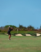 A golfer is captured mid-swing on a lush green golf course. In the background, there are sand bunkers and some shrubbery, with a clear blue sky above.