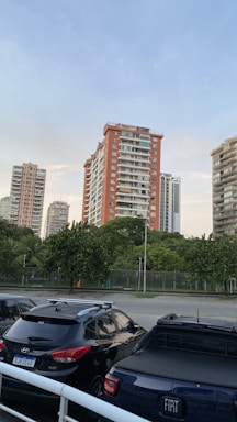 Several high-rise residential buildings surrounded by lush greenery, with parked cars in the foreground. The sky is clear with a few clouds, indicating a calm and peaceful setting.