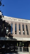 A building facade with a sign reading 'Computer Science Graduate School' in large uppercase letters. The structure is made of reddish-brown bricks with vertical windows. The sky is clear and blue, and there are some shadows from trees visible on the building.