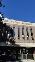 A building facade with a sign reading 'Computer Science Graduate School' in large uppercase letters. The structure is made of reddish-brown bricks with vertical windows. The sky is clear and blue, and there are some shadows from trees visible on the building.