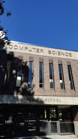 A building facade with a sign reading 'Computer Science Graduate School' in large uppercase letters. The structure is made of reddish-brown bricks with vertical windows. The sky is clear and blue, and there are some shadows from trees visible on the building.