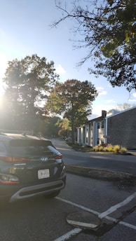 A residential street scene with a parked Hyundai Kona car in the foreground. The sun is shining brightly from the left, casting long shadows and illuminating the trees lining the street. The buildings along the road are a mix of modern and traditional styles, and the sky is clear with a few clouds.