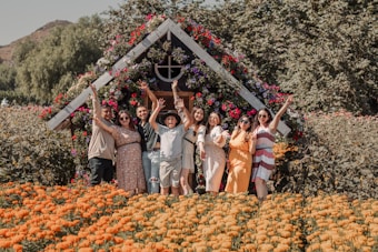 A group of people are posing happily in front of a small, flower-covered structure. The backdrop consists of a lush, colorful garden with orange marigold flowers in the foreground. Each person displays a joyful expression, with some raising their arms in celebration.
