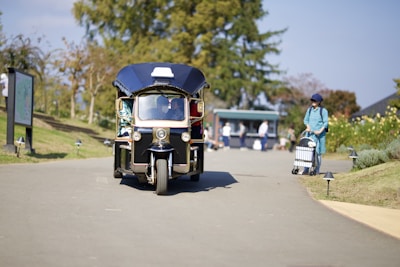 A three-wheeled vehicle, resembling a tuk-tuk, travels on a paved path in a park-like setting. To the right, a person wearing a blue outfit and hat pushes a cart. Trees and a kiosk are visible in the background.