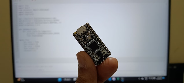 A hand holding a small microcontroller board with a USB port at the top. The background features a computer monitor displaying code or text in a programming environment.