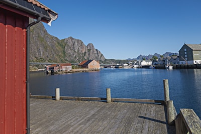 A picturesque harbor scene features a calm body of water surrounded by traditional Scandinavian-style buildings and dramatic rocky mountains in the background. The foreground includes a wooden deck and a red building with a sloped roof. The sky is clear and blue, adding to the serene atmosphere.