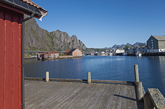 A picturesque harbor scene features a calm body of water surrounded by traditional Scandinavian-style buildings and dramatic rocky mountains in the background. The foreground includes a wooden deck and a red building with a sloped roof. The sky is clear and blue, adding to the serene atmosphere.