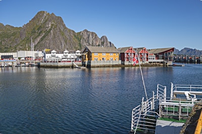 A picturesque waterfront scene features colorful buildings alongside a calm body of water. A large mountain rises prominently in the background, and a dock with railings is visible in the foreground. A Norwegian flag is seen on the dock.