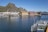 A picturesque waterfront scene features colorful buildings alongside a calm body of water. A large mountain rises prominently in the background, and a dock with railings is visible in the foreground. A Norwegian flag is seen on the dock.