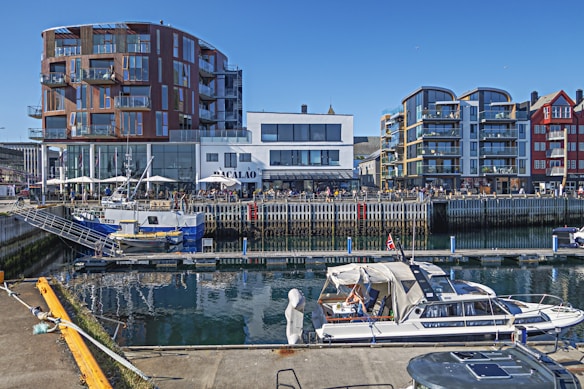 A vibrant harbor scene with modern buildings lining the waterfront. Several boats are moored at the docks in the foreground, including a small fishing vessel and a pleasure yacht. The buildings have a contemporary architectural style with large windows and balconies. People can be seen enjoying the outdoor seating area of a restaurant named Bacalao.