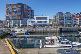 A vibrant harbor scene with modern buildings lining the waterfront. Several boats are moored at the docks in the foreground, including a small fishing vessel and a pleasure yacht. The buildings have a contemporary architectural style with large windows and balconies. People can be seen enjoying the outdoor seating area of a restaurant named Bacalao.