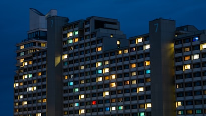 A large residential apartment building with a grid-like pattern of windows. Many windows are illuminated with different colors such as warm yellows, blues, and whites, giving a lively impression. The building is set against a deep blue sky, suggesting nighttime.