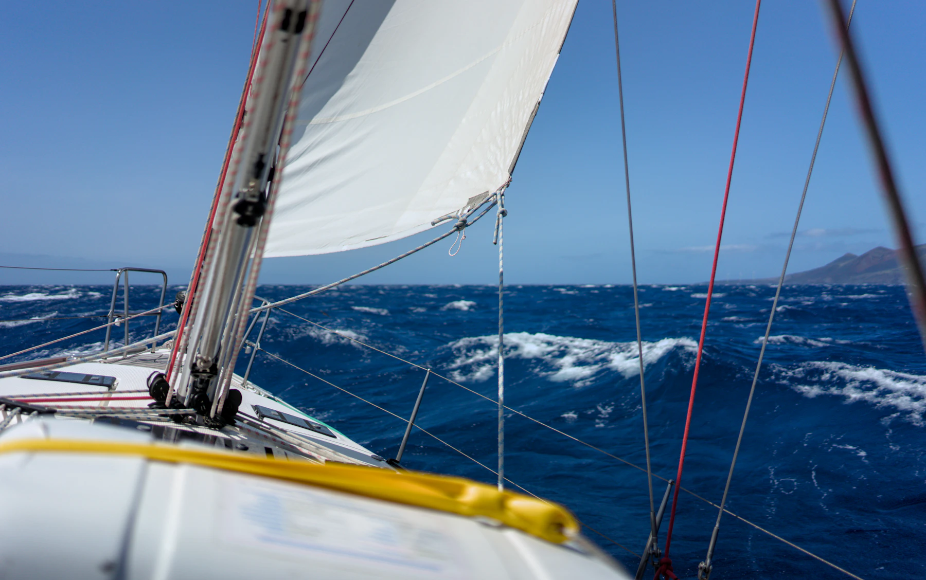 Looking forward from the side deck of a sailing boat under way and heeled over, the headsail full and deep blue water breaking past the rail.